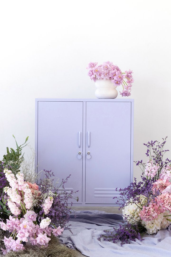 Mustard Made The Midi Locker in lilac with similar coloured flowers lying around it and a lilac blanket underneath